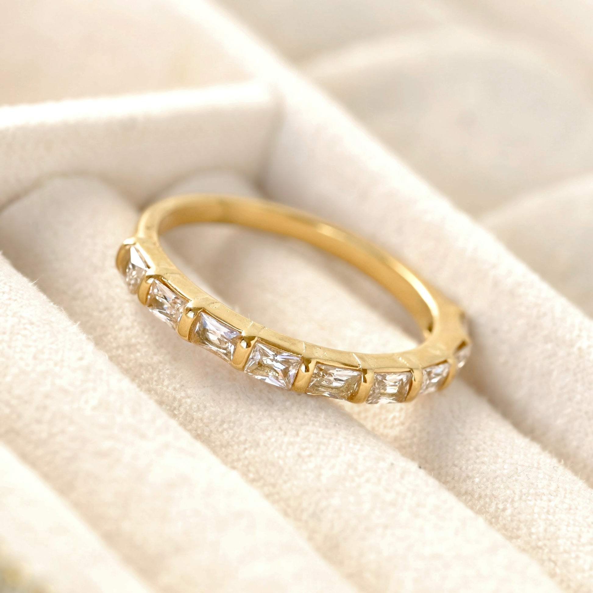 A yellow gold half eternity ring featuring baguette-cut clear stones, resting on a cream-colored velvet ring box.