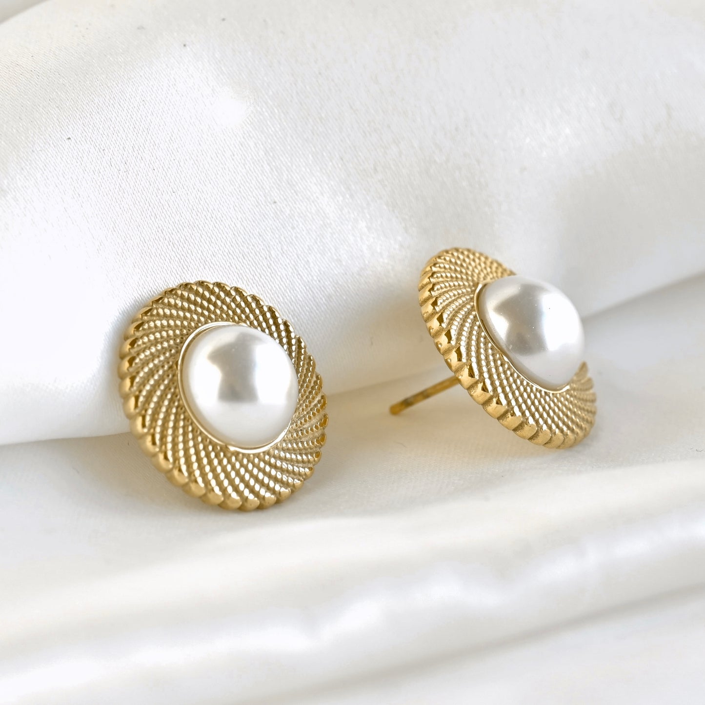 A pair of gold round stud earrings with a textured spiral milgrain border and a central white  simulated pearl, displayed on a soft white fabric background.