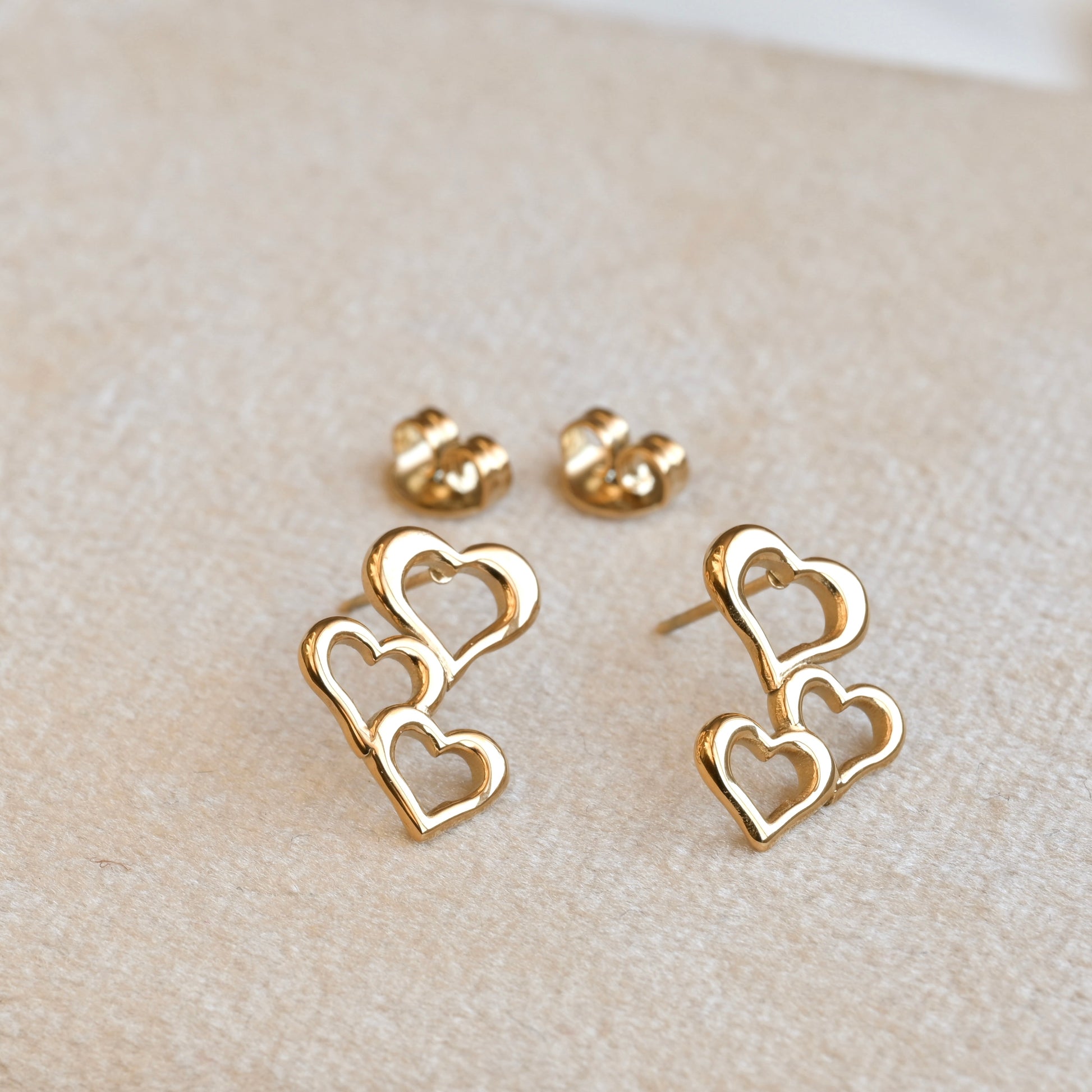 Gold heart-shaped earrings with their butterfly backs on a beige surface