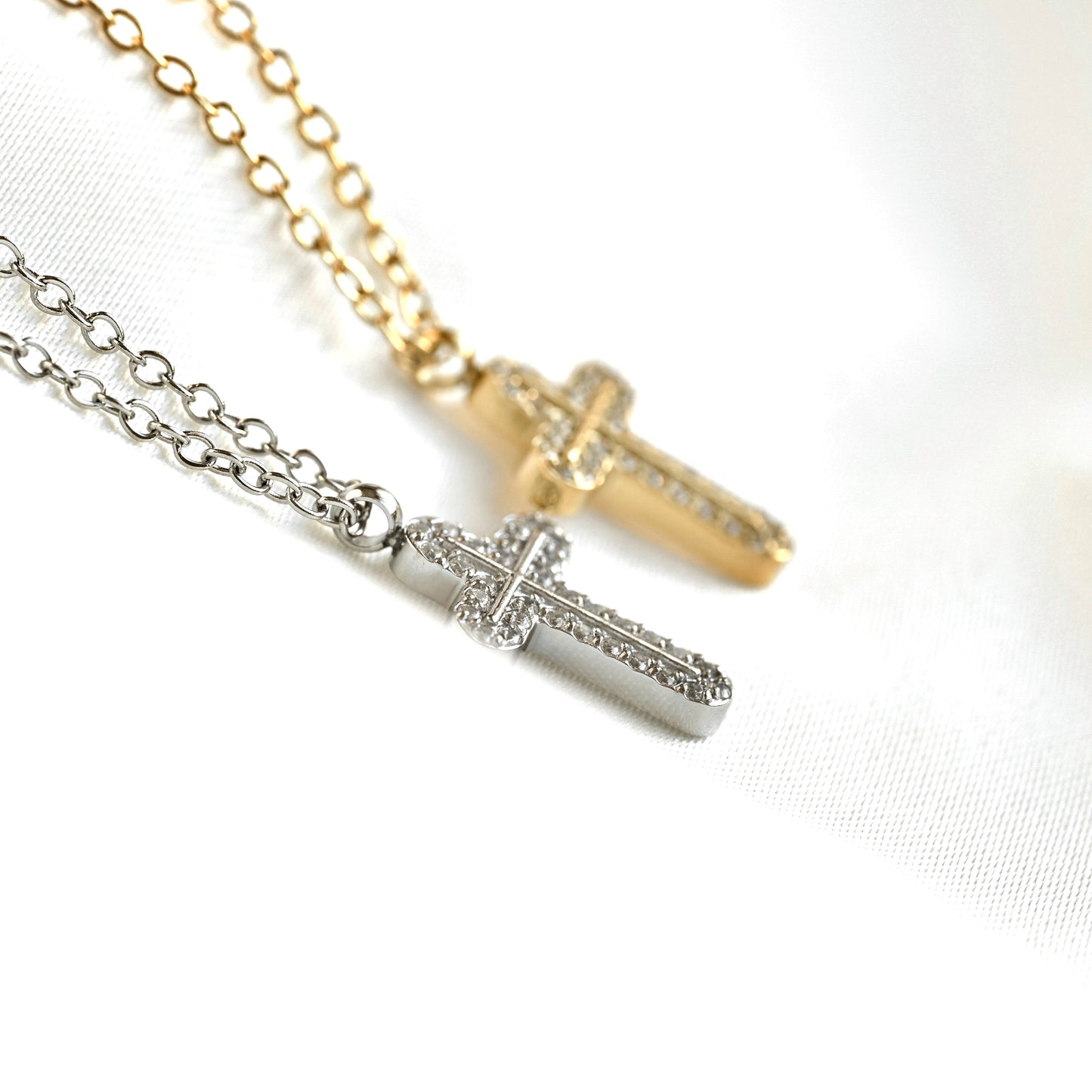 Side close up view of two cross pendant necklaces on  cable chains, one in gold and one in silver, both adorned with small clear stones, displayed on a white textured fabric background.