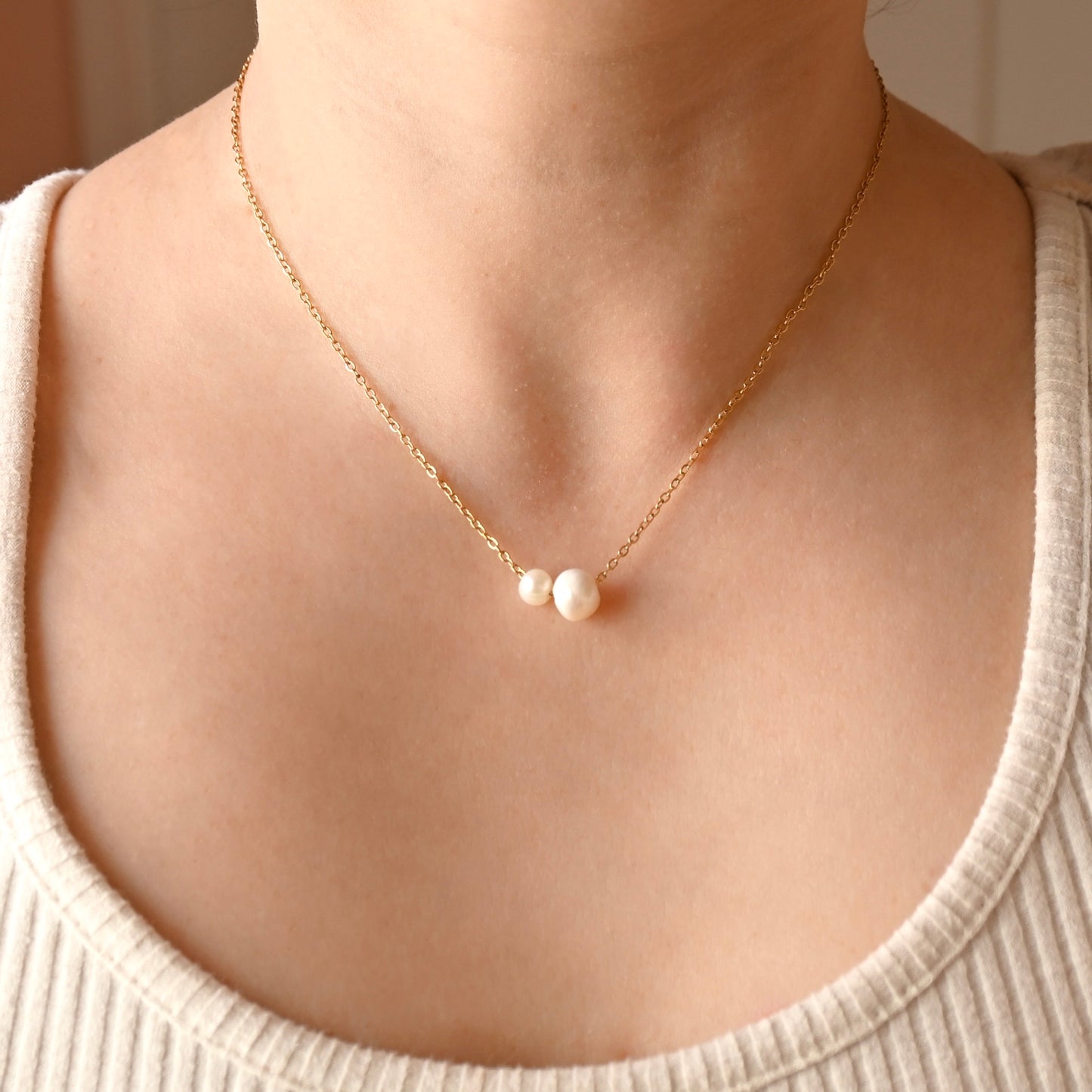 A delicate gold chain necklace with two small white pearl beads at the center, worn on a model's neck over a ribbed cream-colored top.