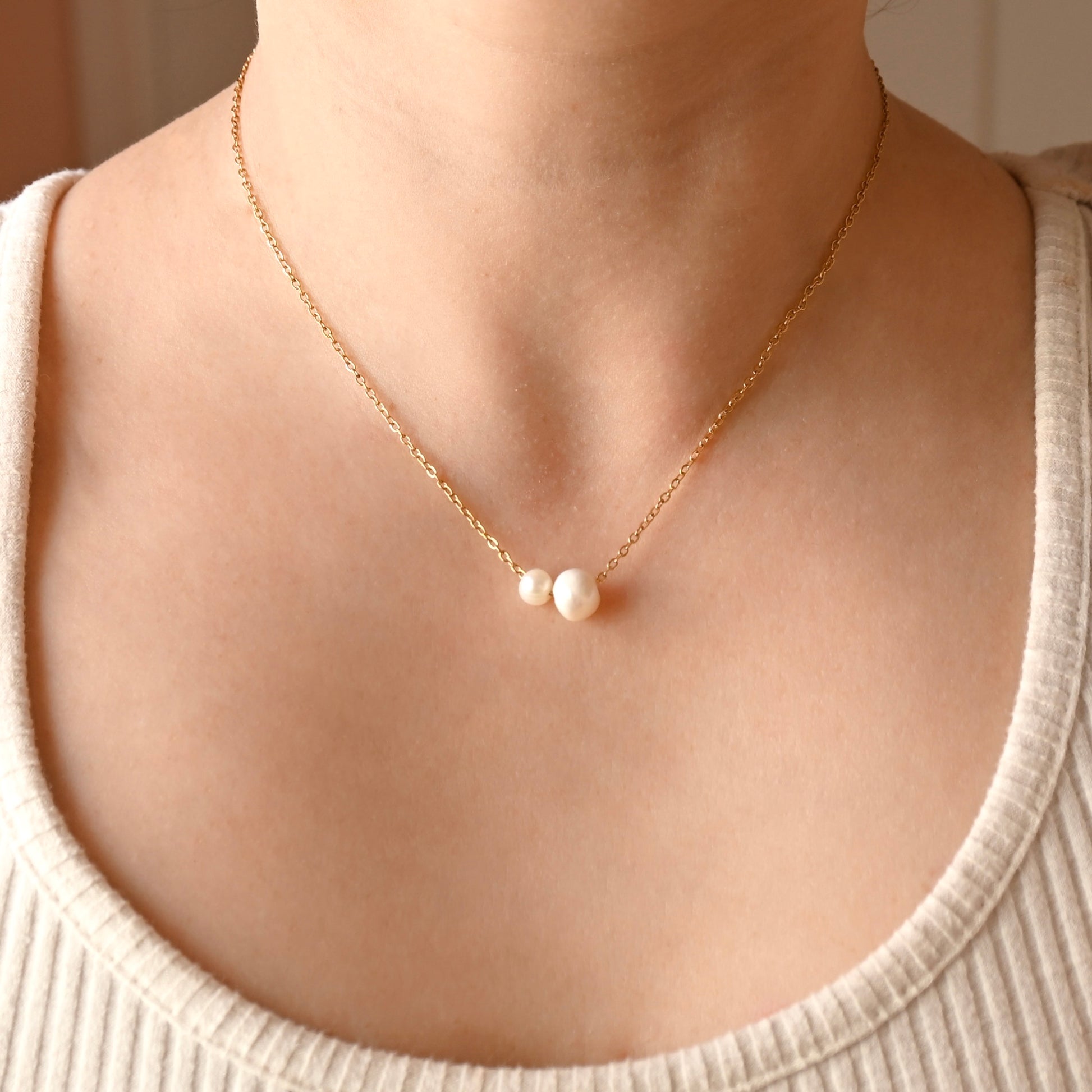 A delicate gold chain necklace with two small white pearl beads at the center, worn on a model's neck over a ribbed cream-colored top.