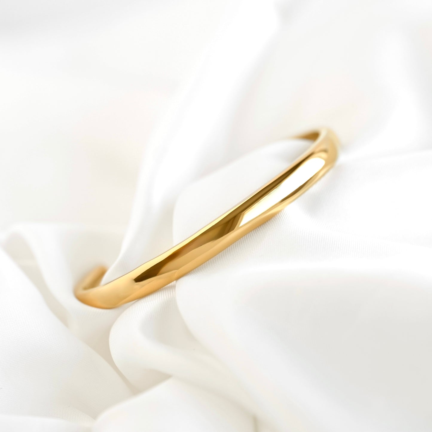 A polished gold dome cuff bracelet resting on draped white satin fabric.