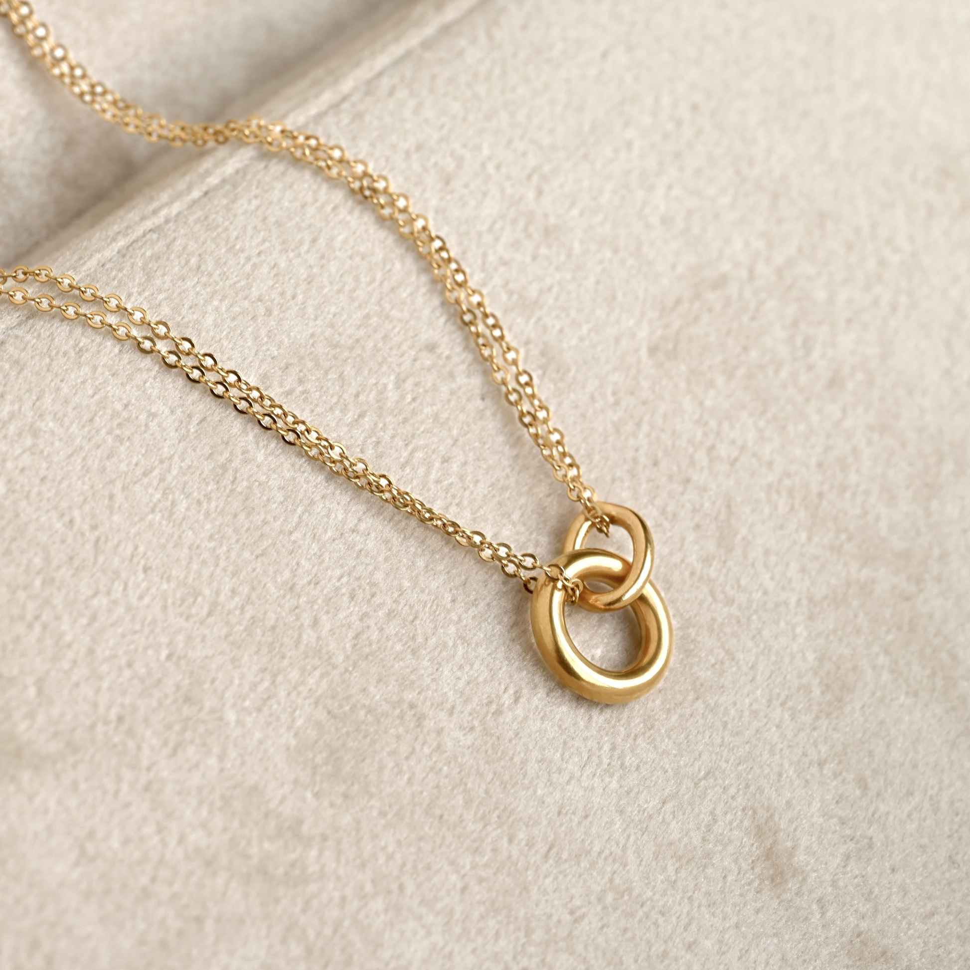 A gold chain necklace with a polished interlocking double-ring pendant, displayed on a textured beige velvet surface.