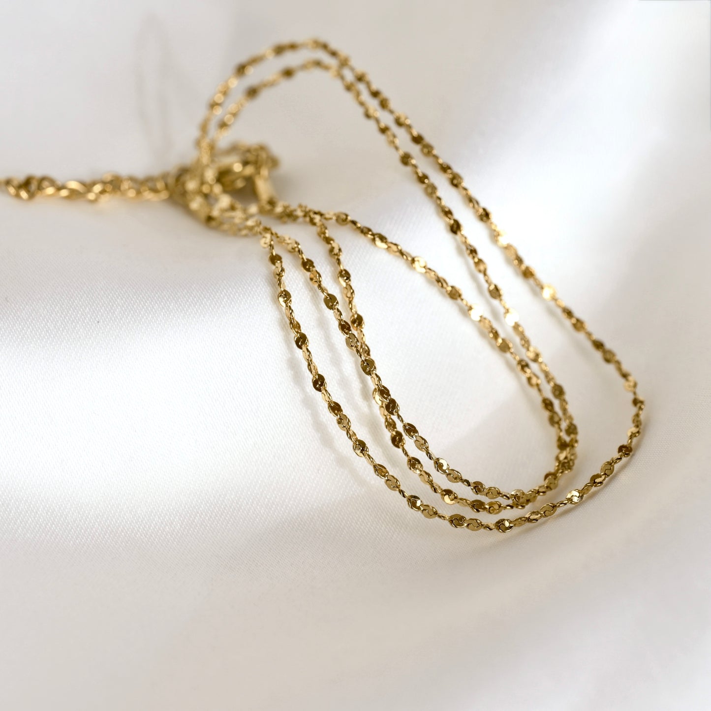 A delicate gold sparkle chain necklace draped in multiple strands on a soft, white textured fabric background.