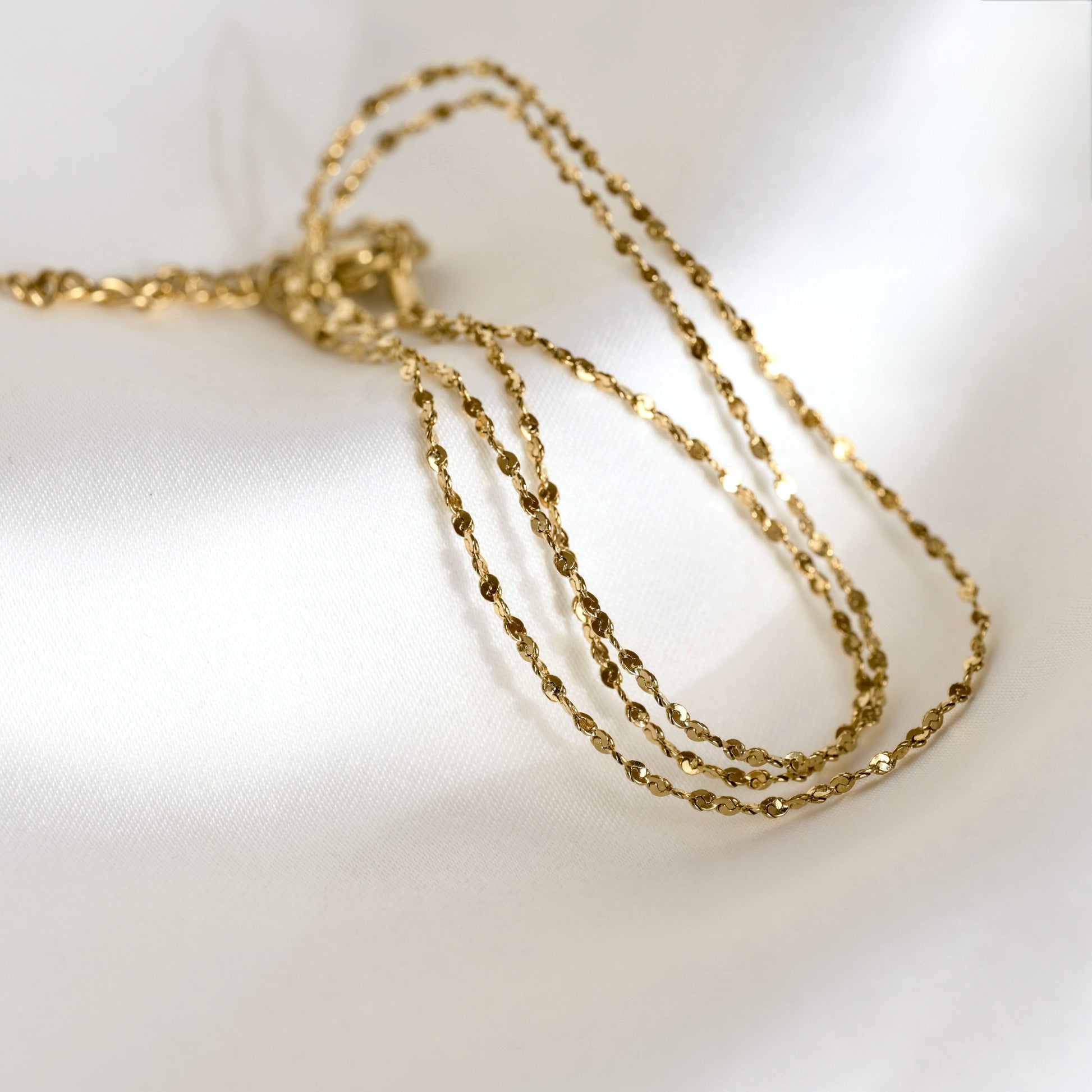 A delicate gold sparkle chain necklace draped in multiple strands on a soft, white textured fabric background.