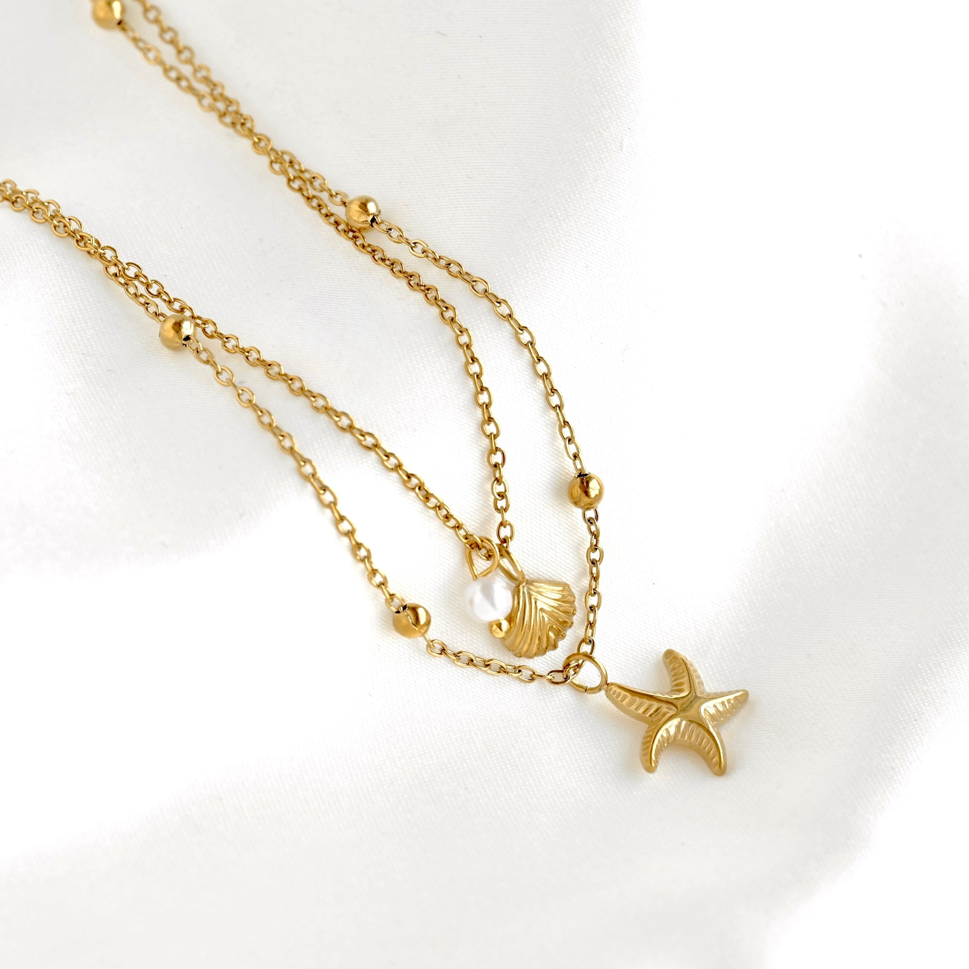 A gold-toned double-strand necklace featuring a small starfish pendant and a seashell charm, displayed on a soft white fabric background.