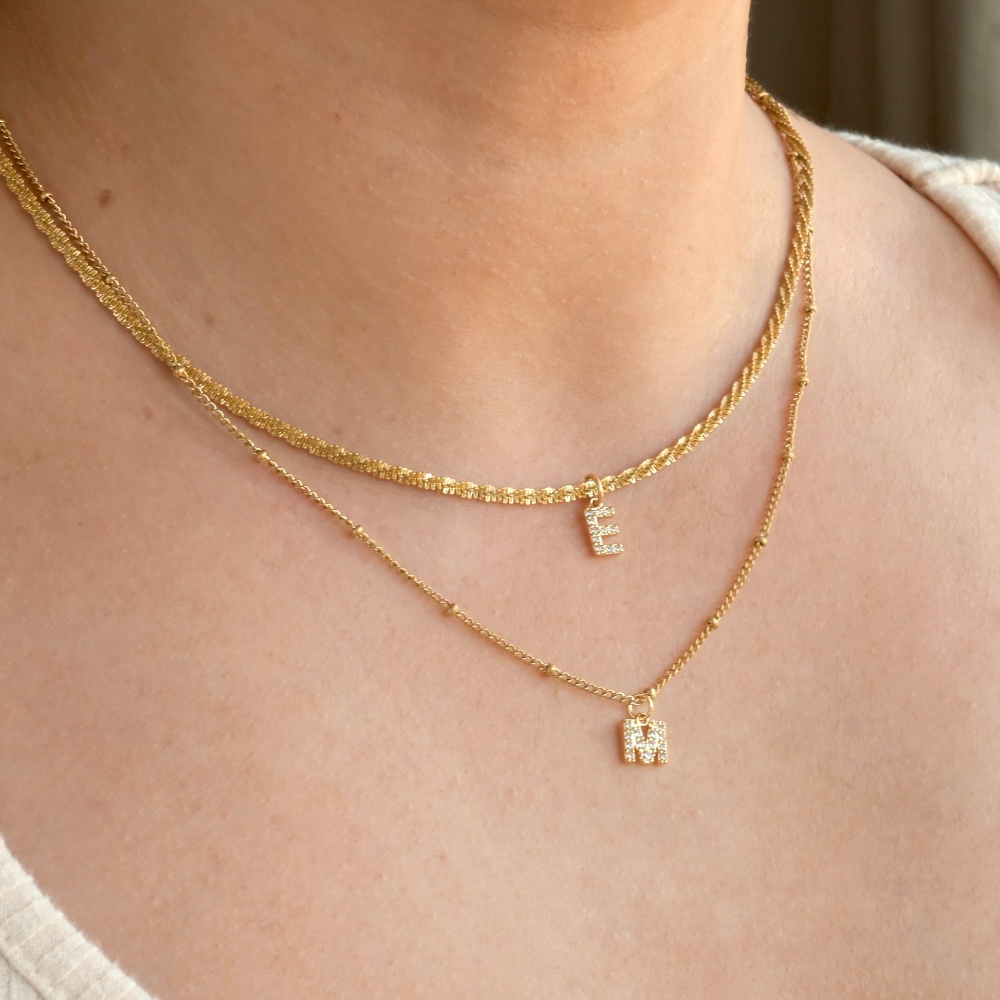 Two gold necklaces layered, one shorter sparkle chain with letter 'E', and one longer satellite chain with letter 'M' on a person's neck