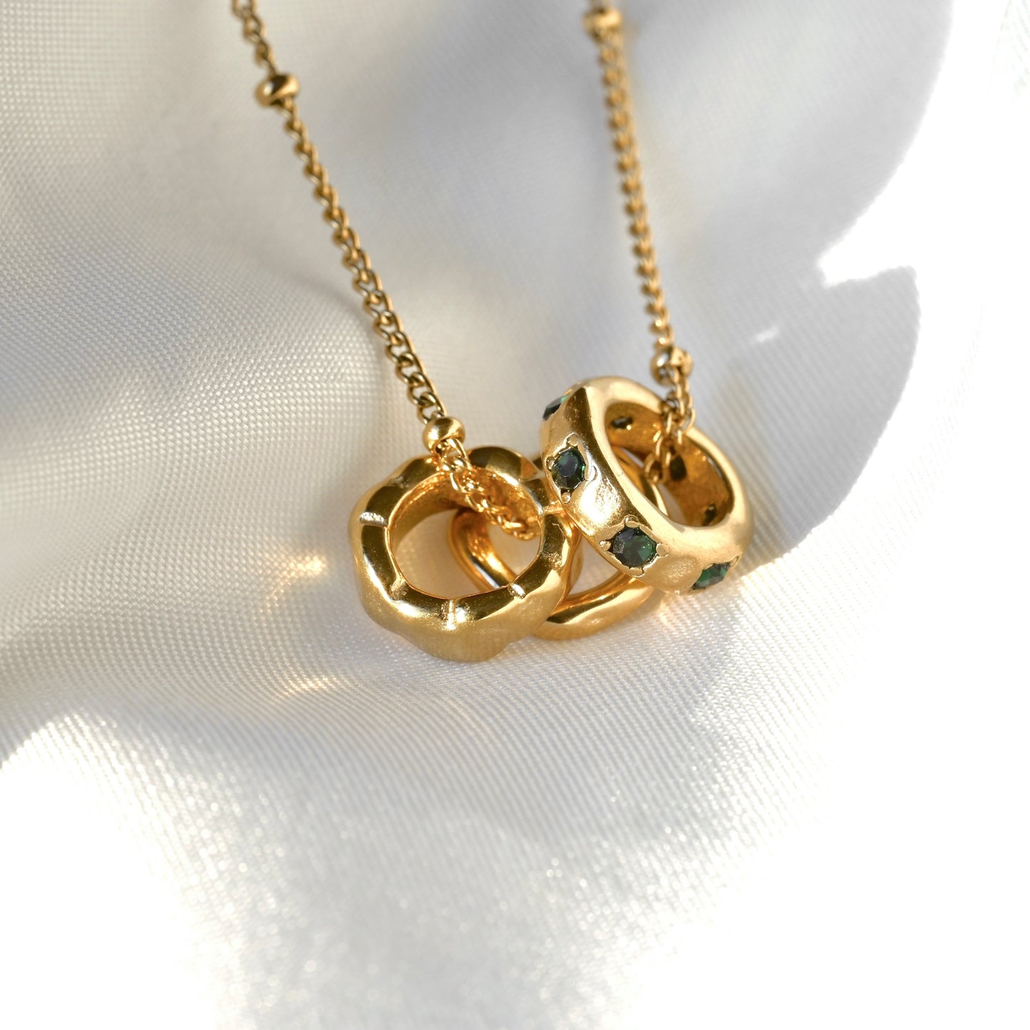 Gold chain necklace with a pendant made of three interlocking textured rings accented with small dark green gemstones, displayed on a white textured fabric background.