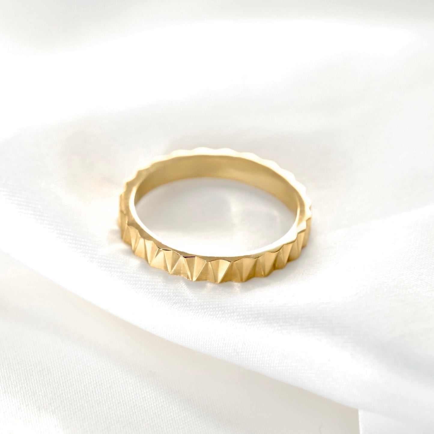 A gold ring with a textured, faceted triangular pattern around the outer edge, resting on a soft, draped white fabric background.