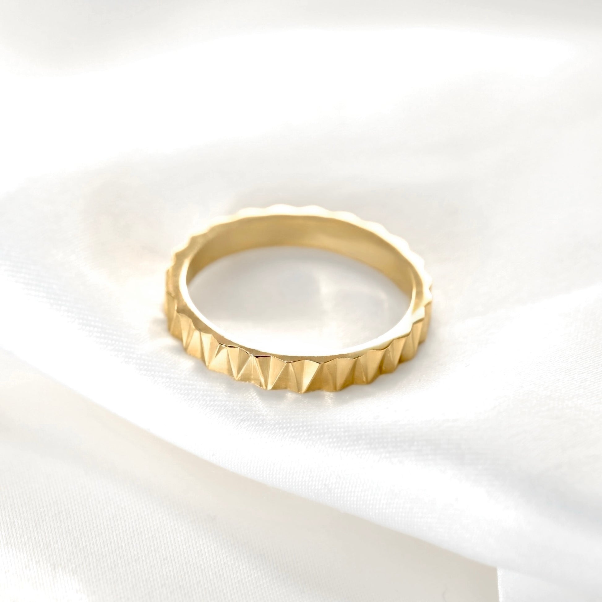 A gold ring with a textured, faceted triangular pattern around the outer edge, resting on a soft, draped white fabric background.