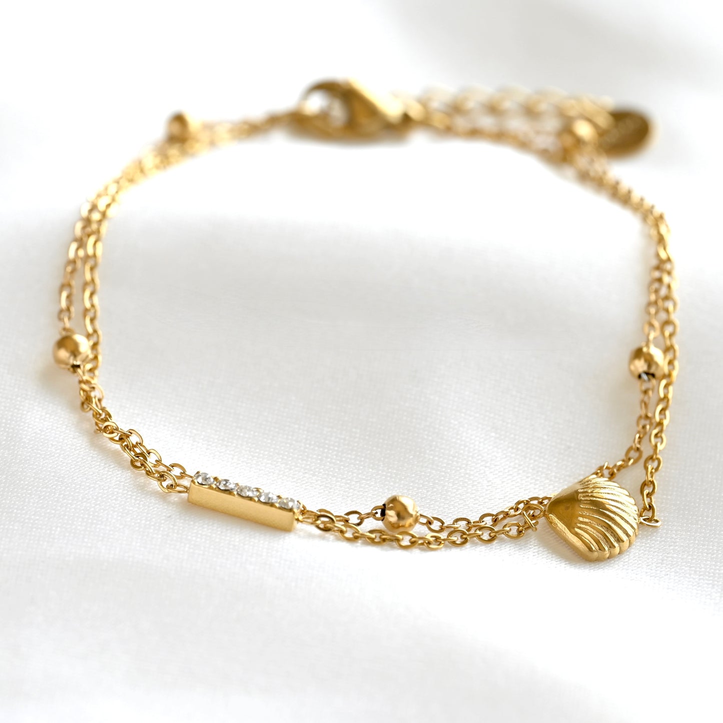 A delicate gold chain bracelet with a lobster clasp, featuring a small seashell charm, a bar with tiny clear stones, and small gold beads, displayed on a white fabric background.