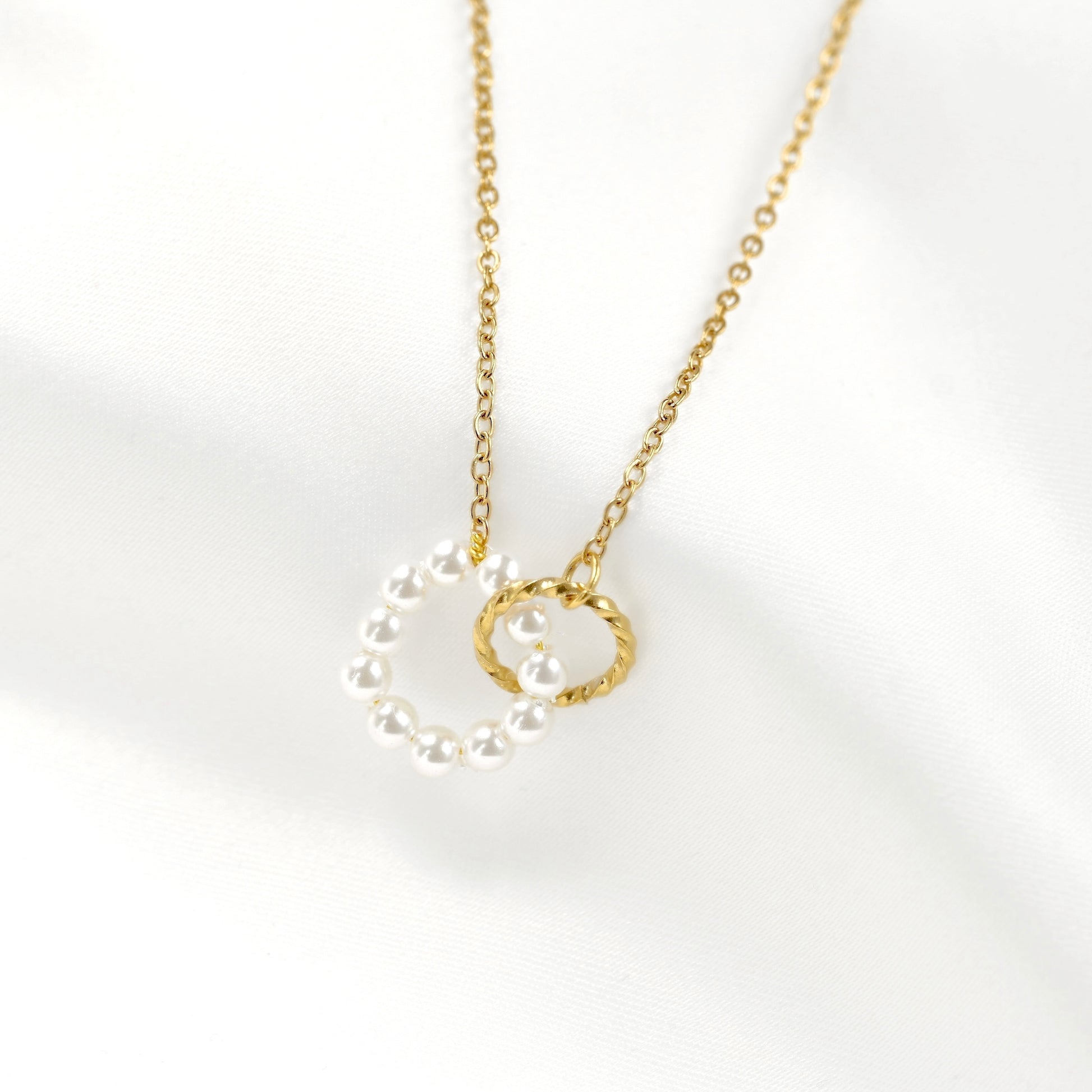 A delicate gold chain necklace featuring a pendant with one open circle made of white pearls and another open twisted gold circle set against a soft white fabric background.