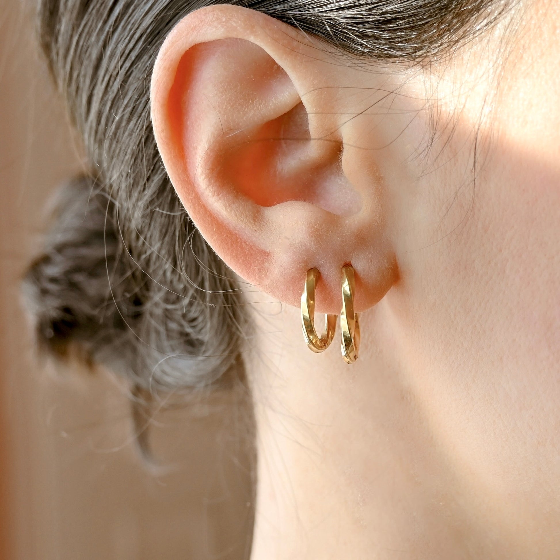 A close-up of a person's ear wearing one small and one bigger gold swirl hoop earrings, with hair pulled back and a soft neutral background.