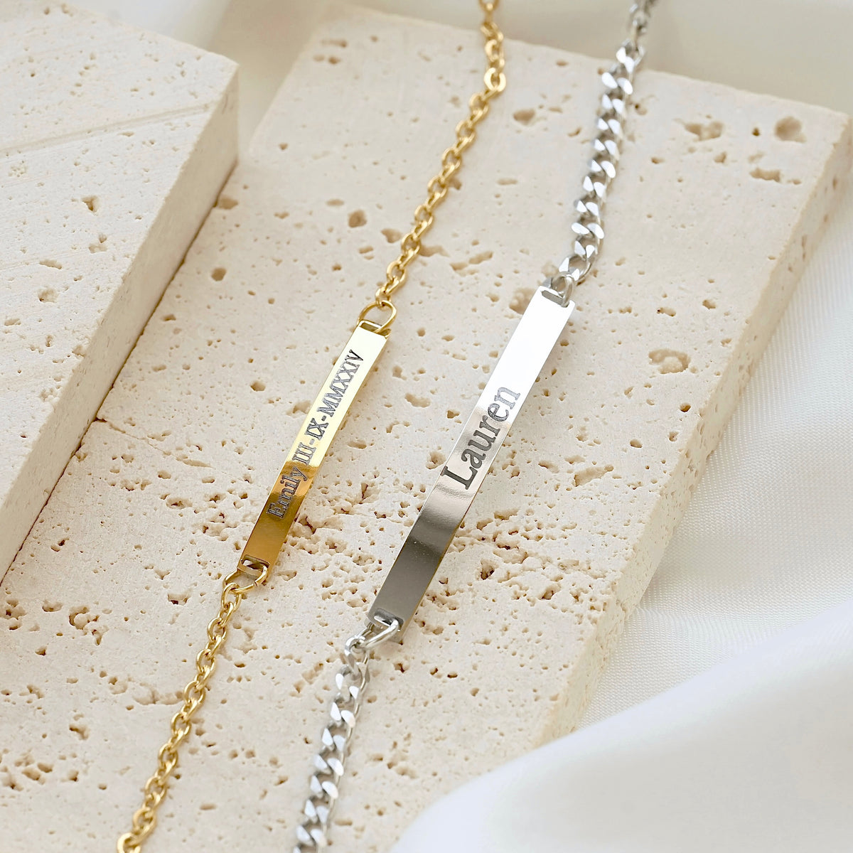 Two personalized nameplates on chains, one gold and one silver, displayed on textured beige stone blocks with a white fabric background. The gold chain reads 'Emily + 01.