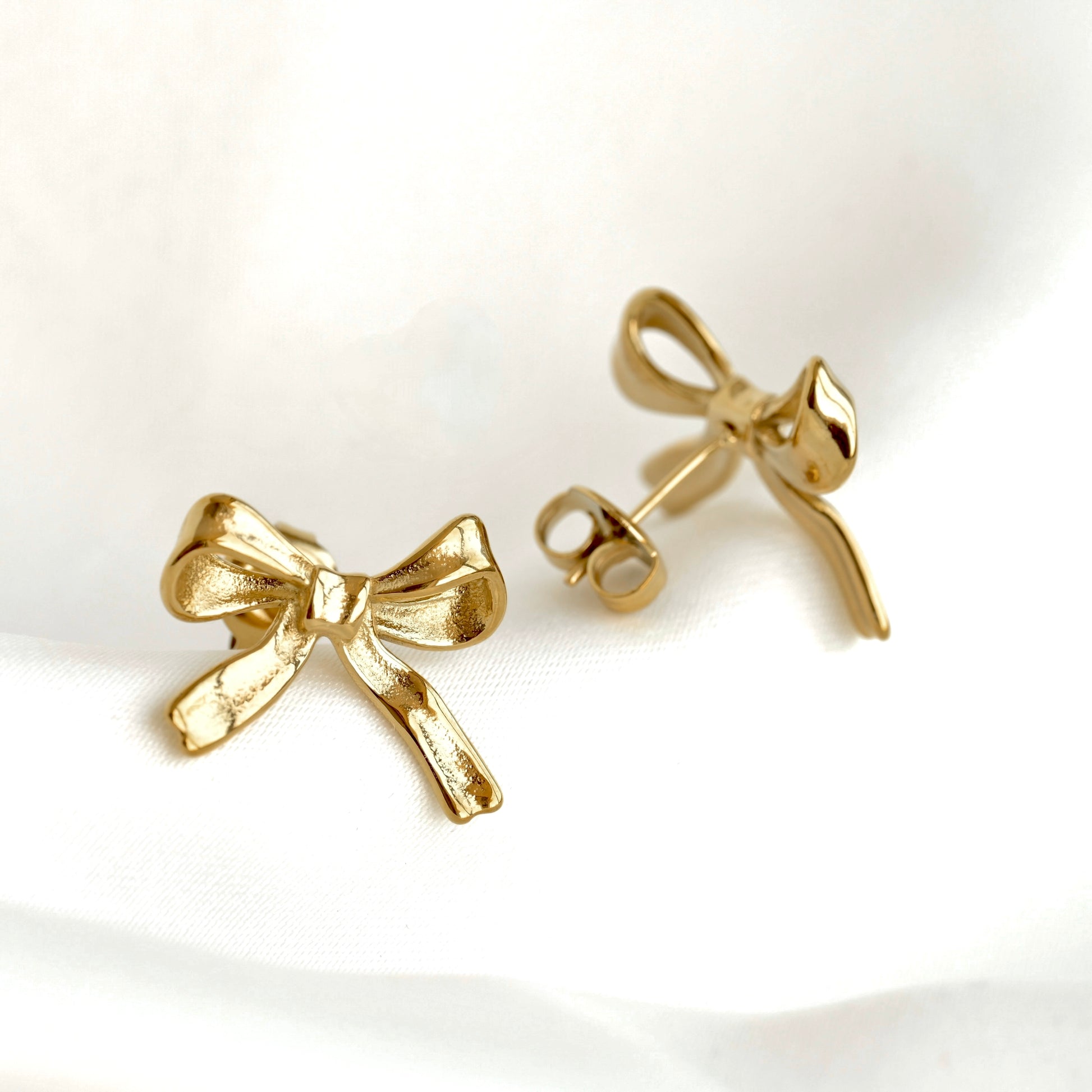 A pair of gold bow stud earrings, with one shown from the front and the other from the side/back to display the post and backing, resting on a white fabric background.