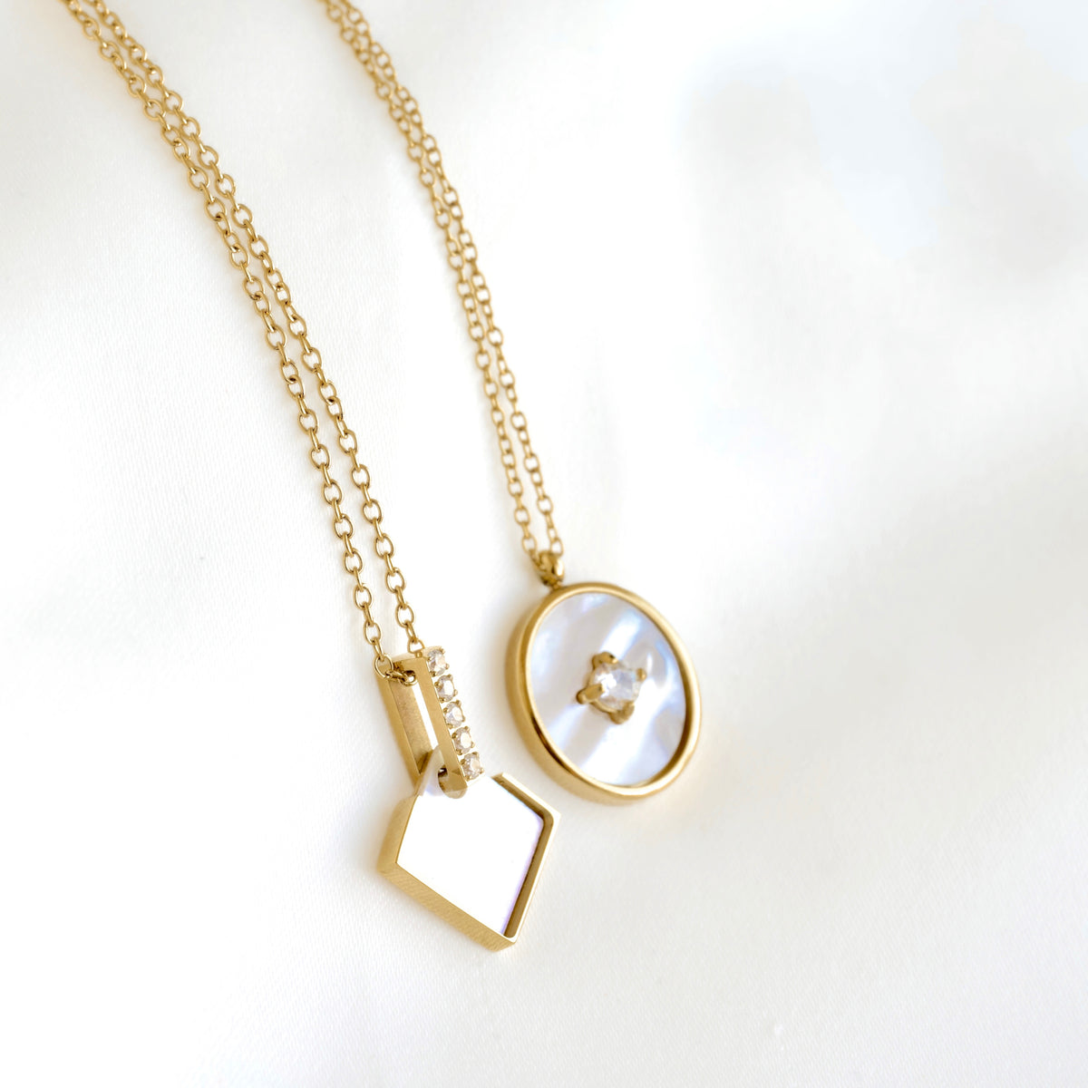 Gold necklace with two pendants: a small geometric gold charm and an oval pendant featuring a white mother-of-pearl inlay and a tiny clear gemstone, displayed on a soft white background.