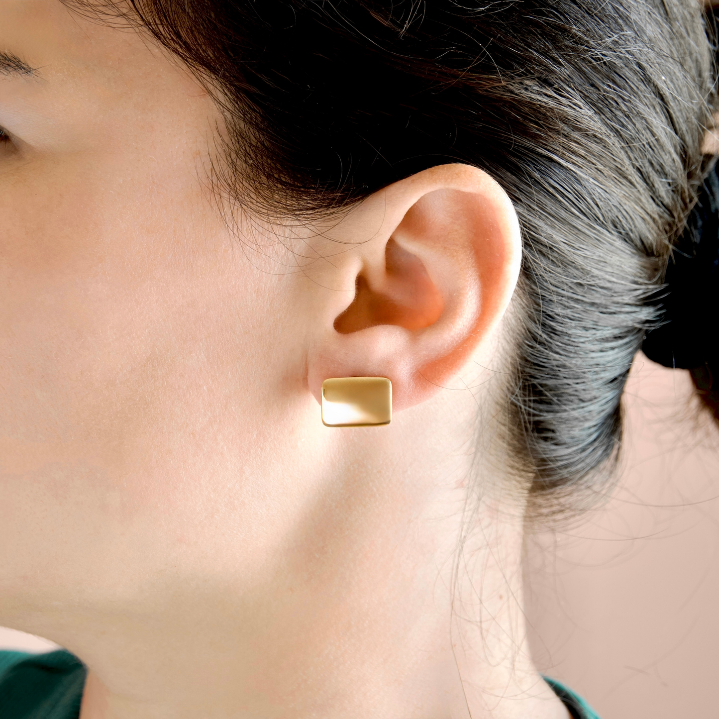 A close-up side profile of a woman wearing a small, rectangular gold stud earring on her earlobe.