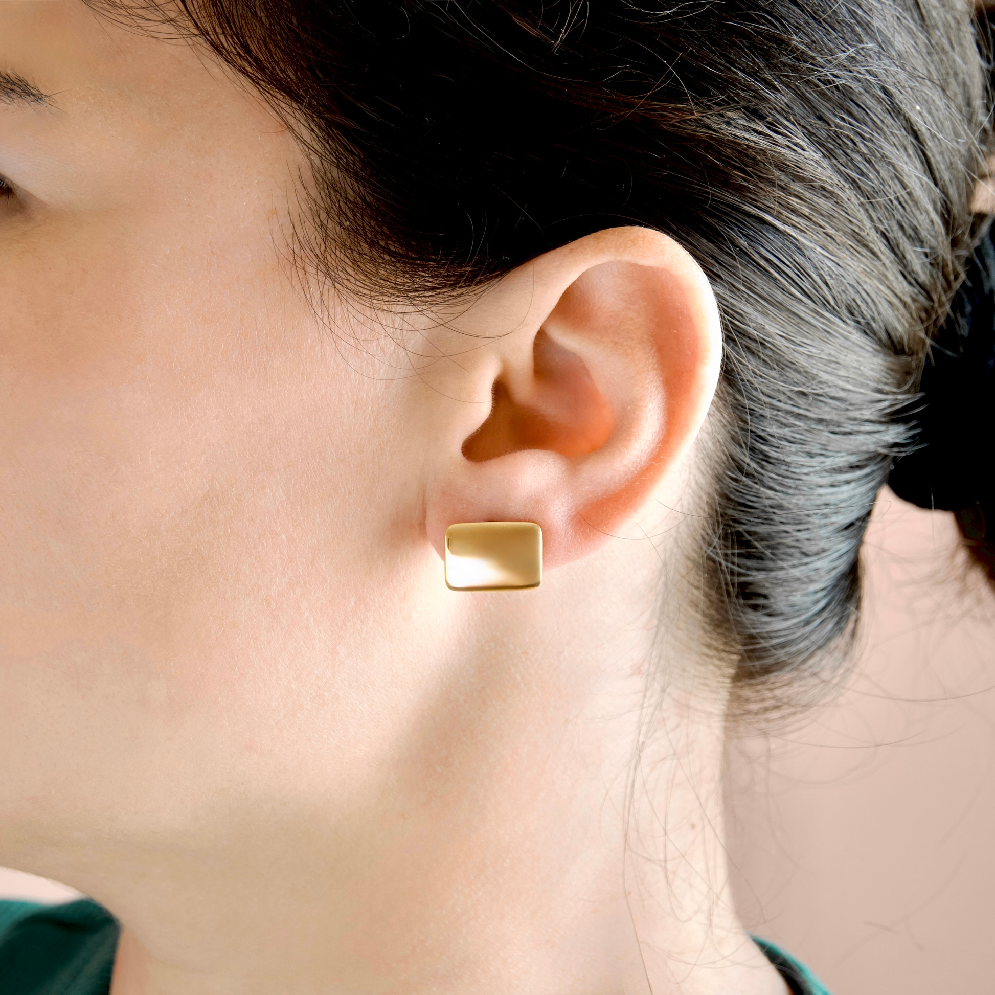 A close-up side profile of a woman wearing a small, rectangular gold stud earring on her earlobe.