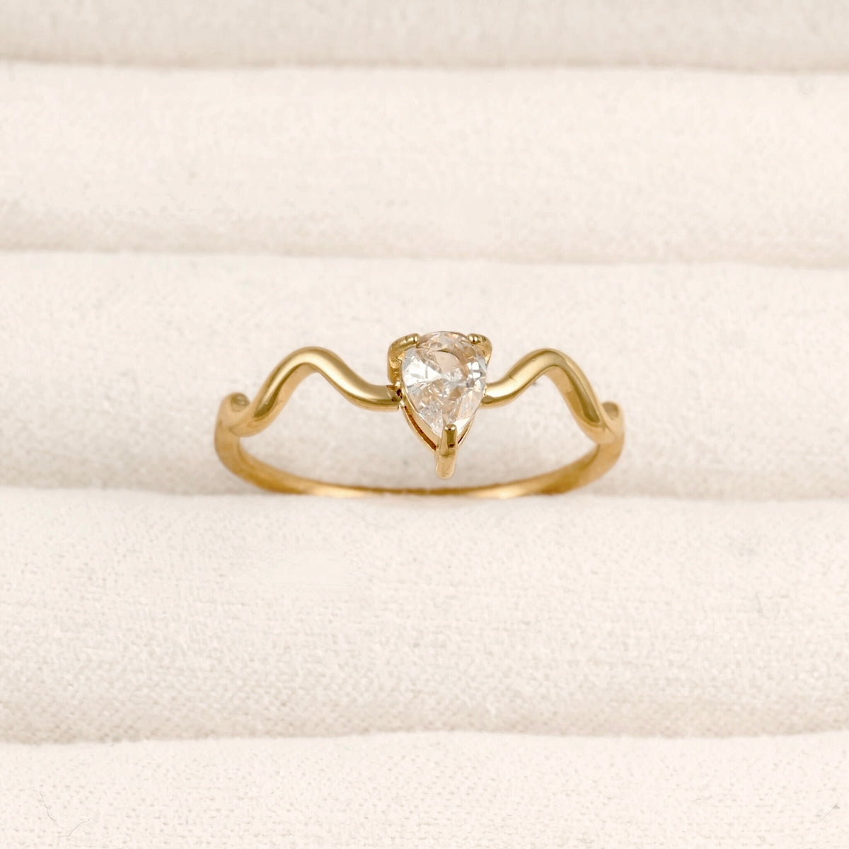 A yellow gold ring with a wavy, open band design and a central pear-cut clear gemstone, displayed on a textured cream-colored ring box.