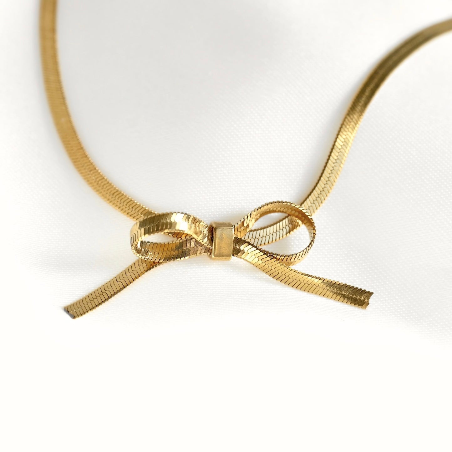 A thin gold herringbone chain necklace featuring a central bow charm, displayed on a clean white background.