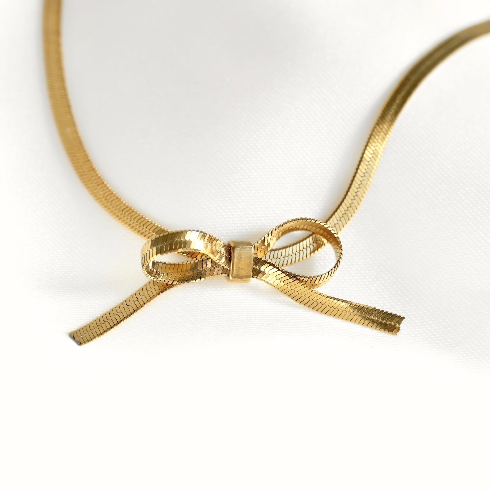 A thin gold herringbone chain necklace featuring a central bow charm, displayed on a clean white background.