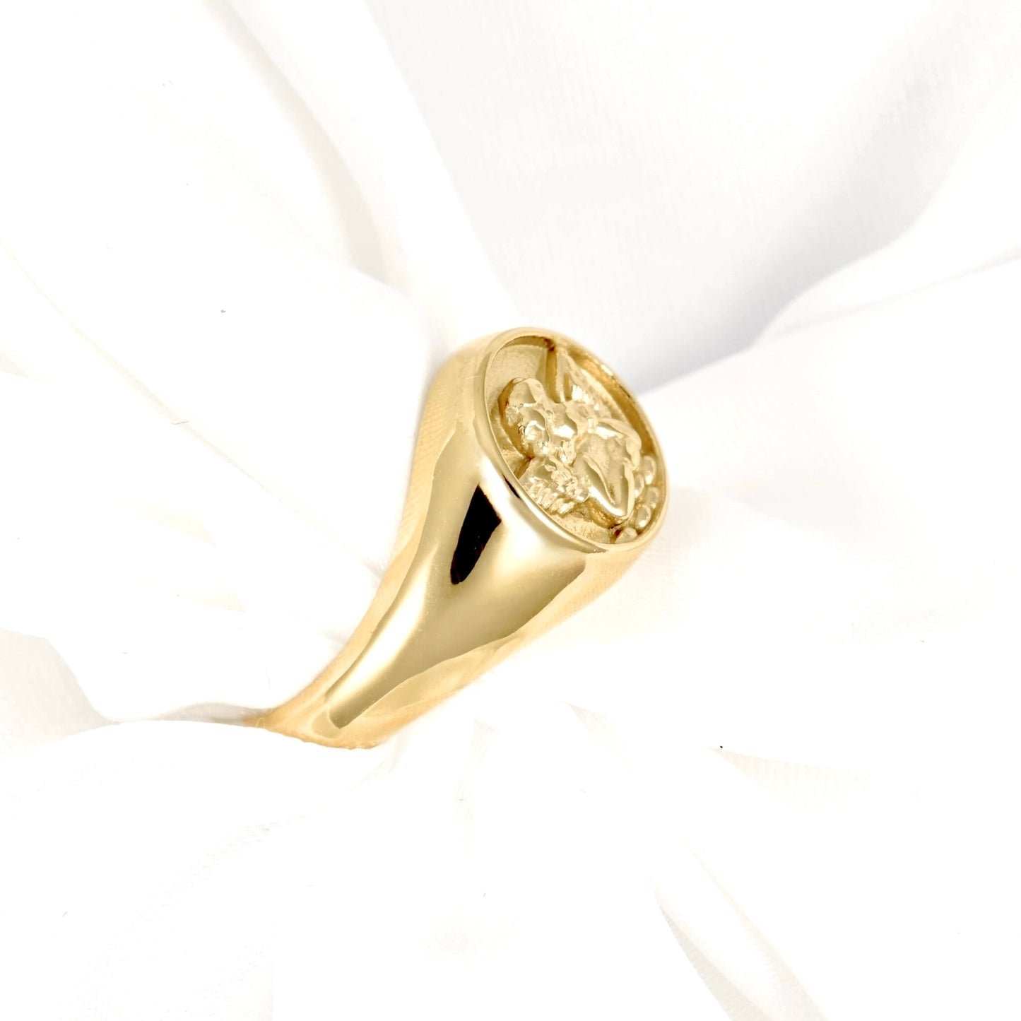 A polished yellow gold signet ring featuring an embossed angel on its oval face, displayed on a soft white fabric background.