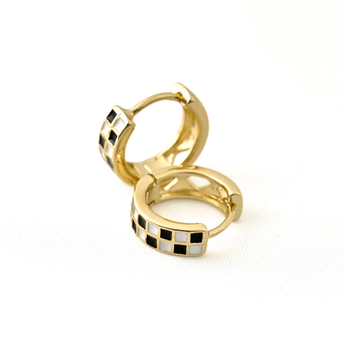 A pair of gold huggie hoop earrings with a checkered black and white enamel pattern on the front, displayed on a clean white background.
