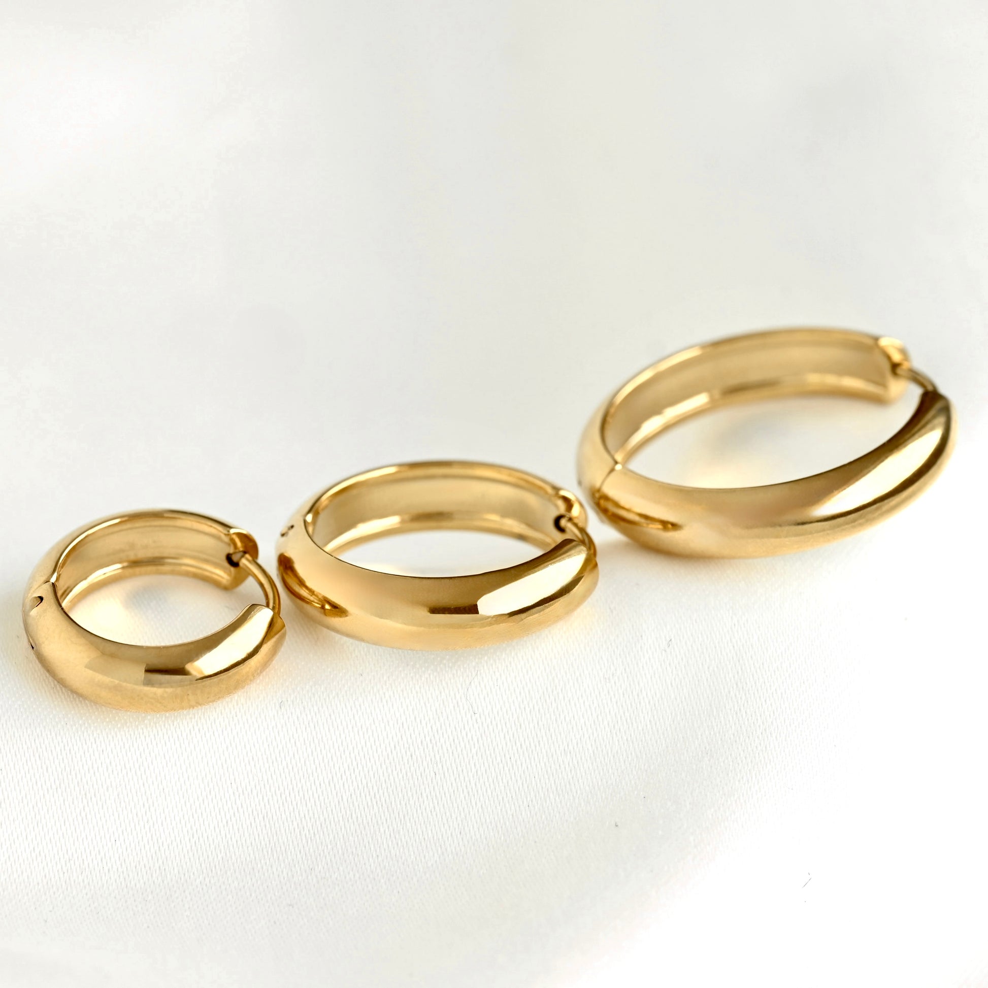 Three gold-plated thick hoop earrings in different sizes, arranged diagonally on a clean white textured fabric background.