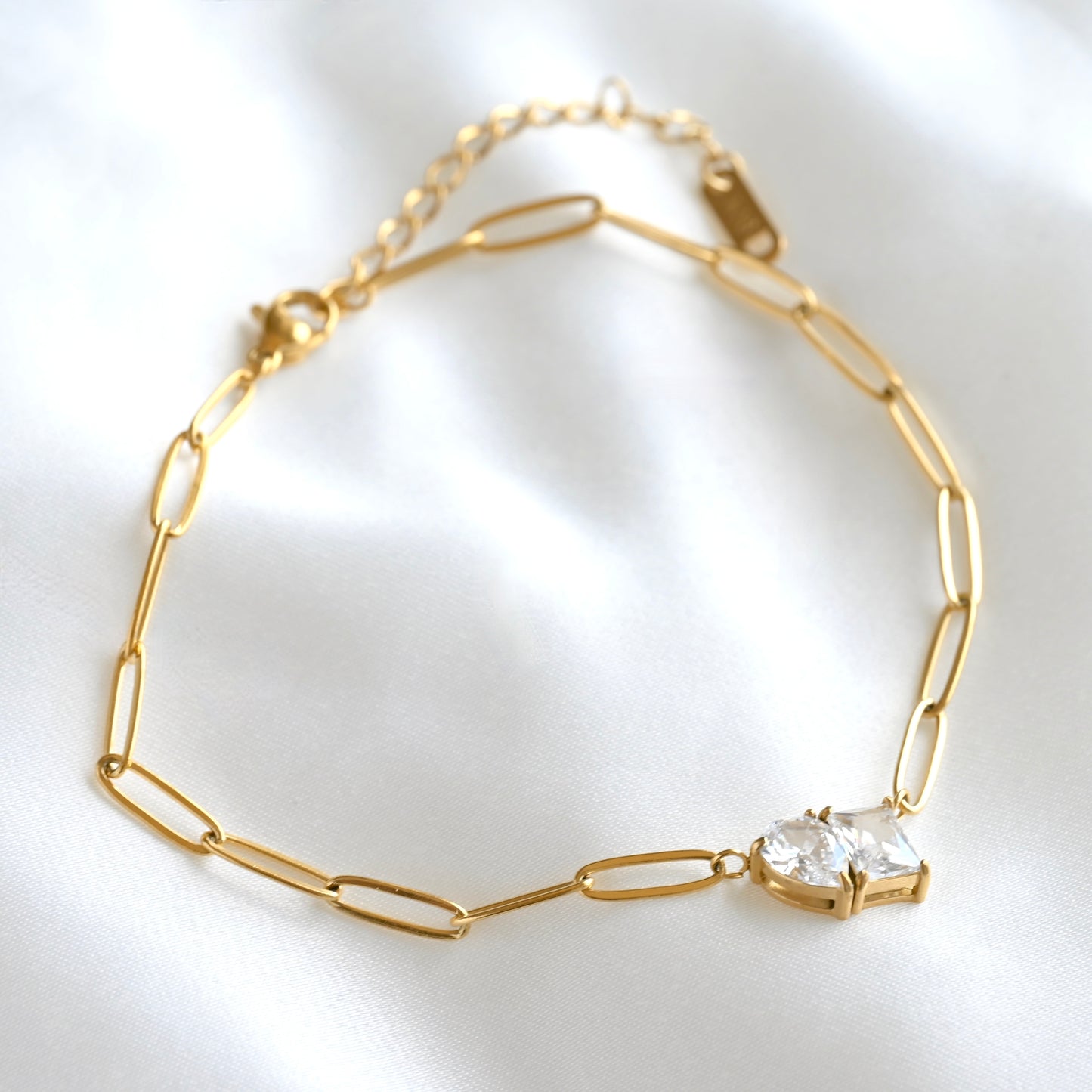 Gold paperclip chain anklet with a clear rectangular  and teardrop stone pendant, displayed on a white satin fabric background.