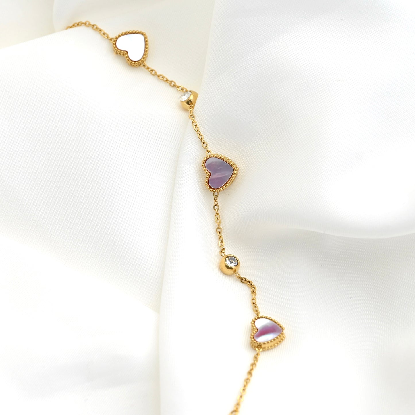 A delicate gold chain bracelet draped on a white fabric background, featuring three heart-shaped charms with shell inlays and two clear bezel set CZ stone accents.