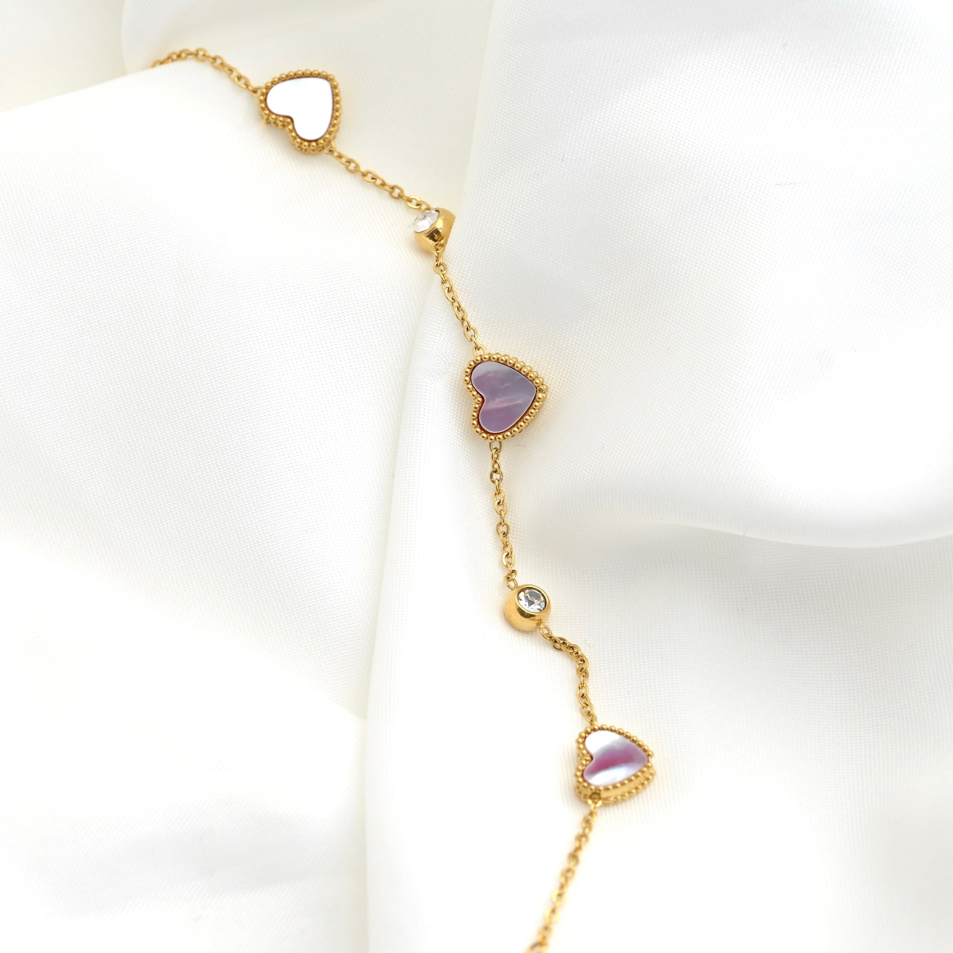 A delicate gold chain bracelet draped on a white fabric background, featuring three heart-shaped charms with shell inlays and two clear bezel set CZ stone accents.