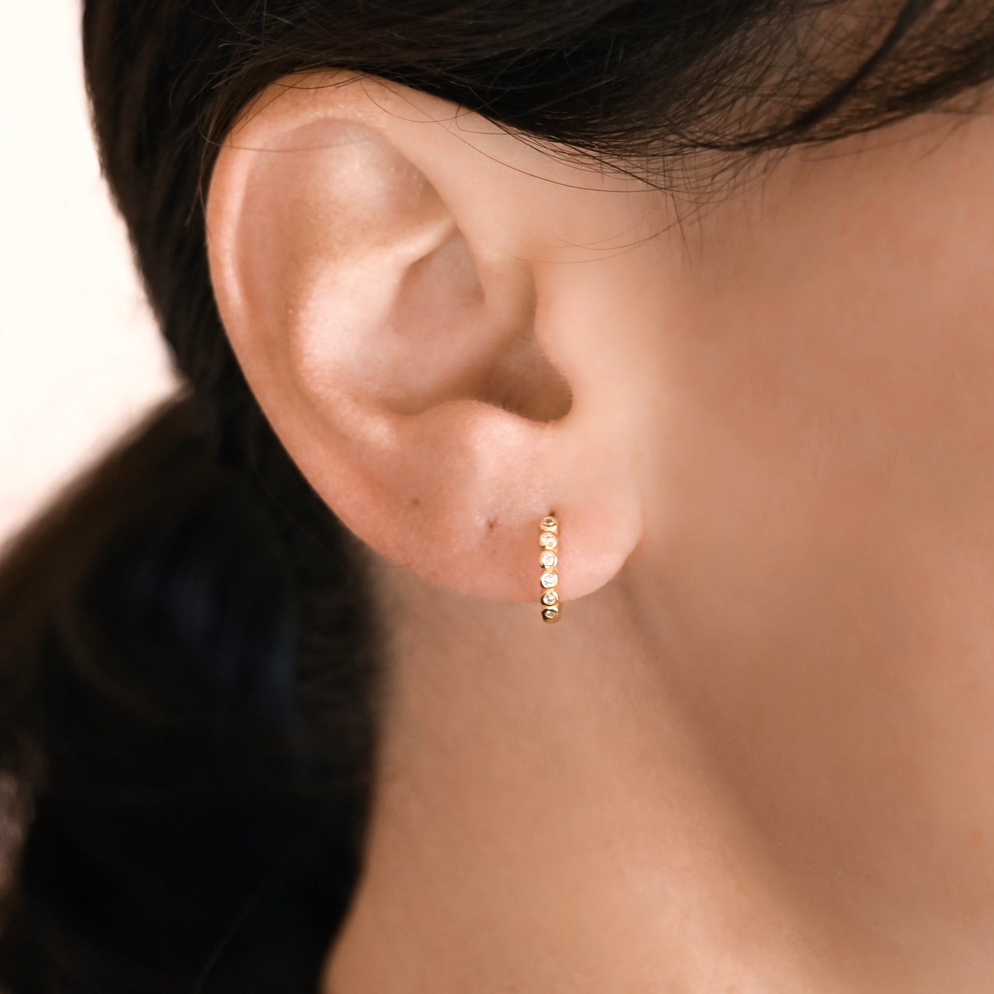 A close-up of a person's ear wearing a small gold huggie hoop earring with a row of tiny round clear stones in a bezel setting, shown against skin and dark hair.