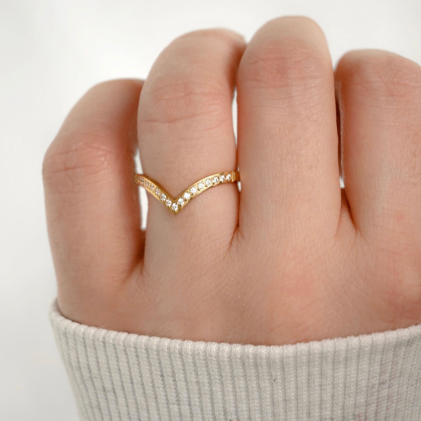 A close-up of a hand wearing a delicate gold V-shaped ring adorned with small clear stones, paired with a light gray ribbed knit sleeve.