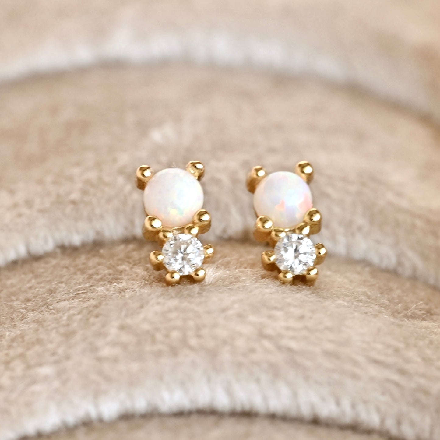 A pair of gold stud earrings featuring a round white opal and a clear round-cut stone, displayed on a soft beige textured fabric background.