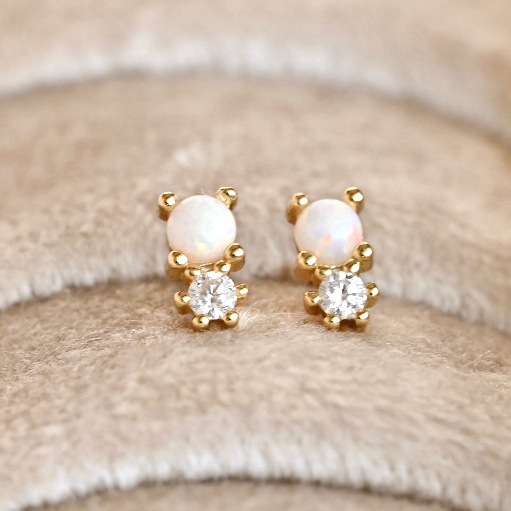 A pair of gold stud earrings featuring a round white opal and a clear round-cut stone, displayed on a soft beige textured fabric background.