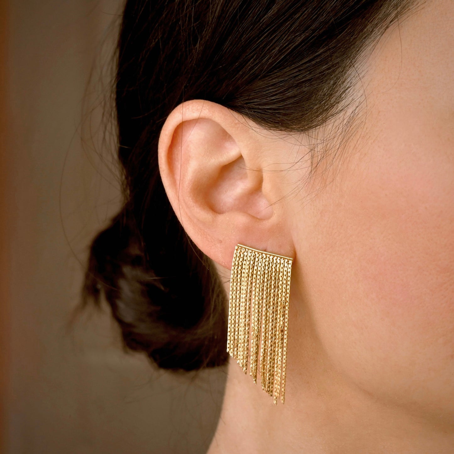 A close-up of a model's ear wearing a gold dangling tassel earring with multiple fine chain strands.