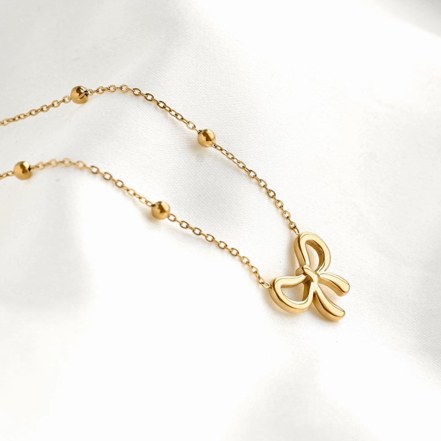 Side view of a delicate gold chain necklace featuring a small bow-shaped pendant and several small gold bead accents, displayed on a soft white fabric background.