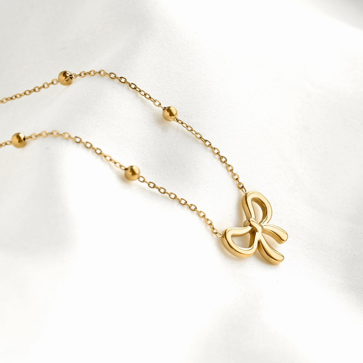 Side view of a delicate gold chain necklace featuring a small bow-shaped pendant and several small gold bead accents, displayed on a soft white fabric background.