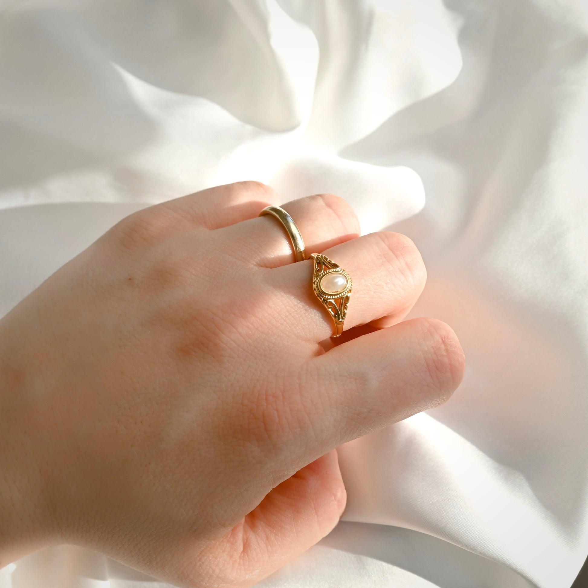 A hand wearing a gold ring with a textured filigree setting and a central oval pearl, posed against a draped white fabric background.