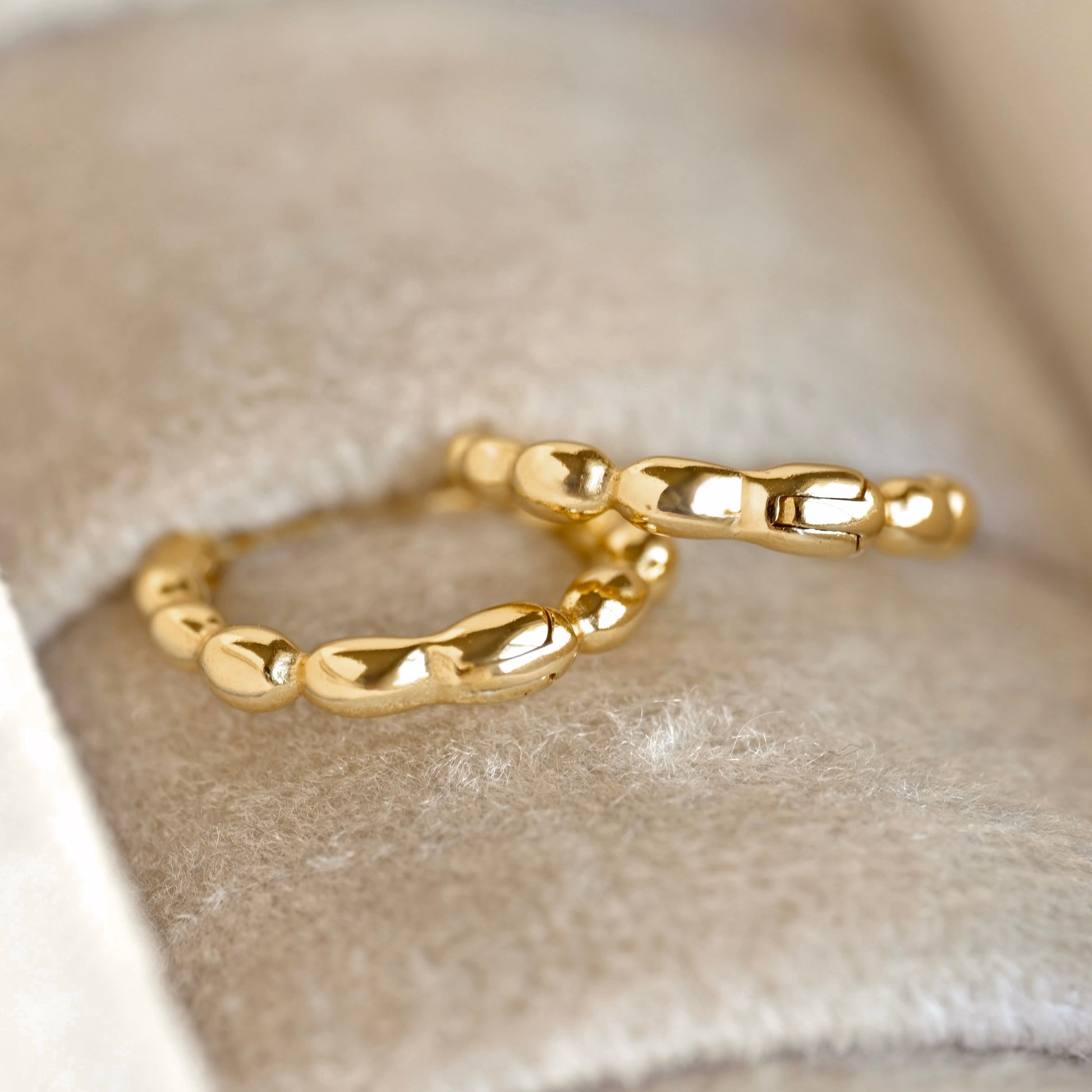 A pair of polished gold beaded hoop earrings resting on a soft beige velvet jewelry box insert.