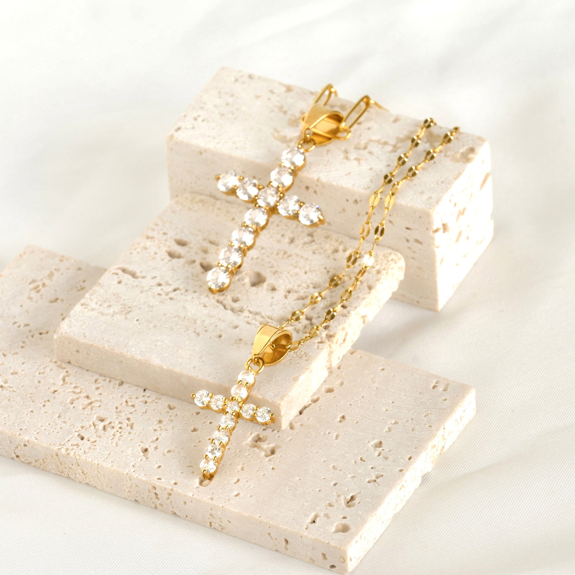 Gold cross necklaces, one medium with a mariner chain and one  large with a paperclip chain on a stone surface