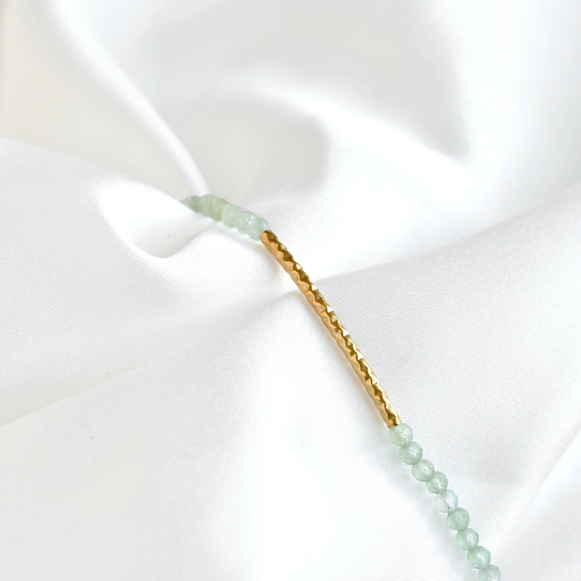 A delicate gold bracelet with light green faceted bead accents and round hammered bar in 18k gold PVD stainless steel displayed on a draped white satin fabric.