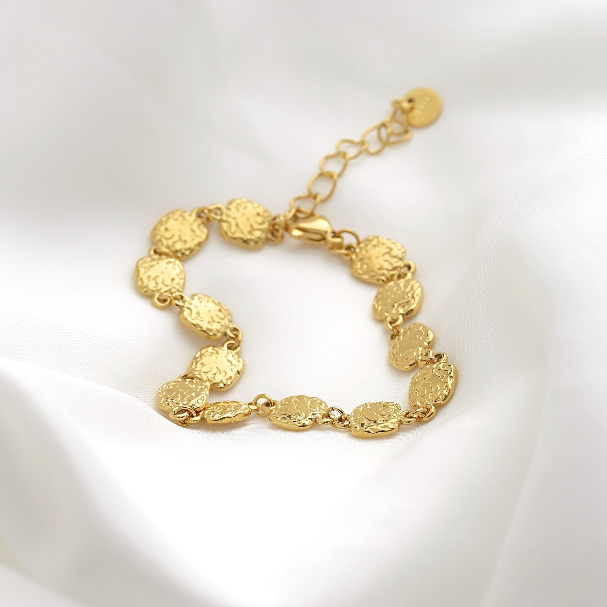 A gold-plated bracelet with textured, disc-like oval links, displayed on a soft white fabric background.