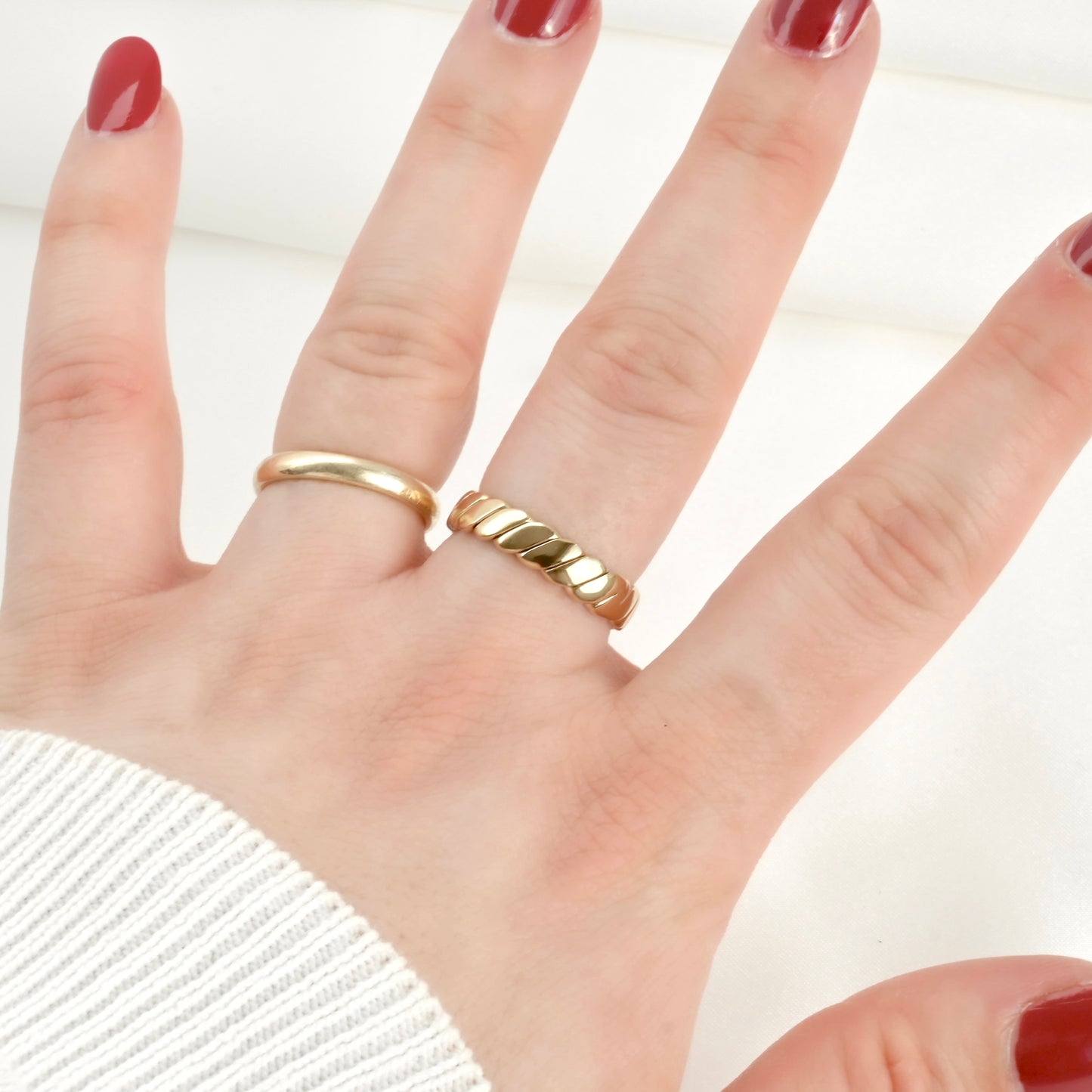 Open hand pose with red nail polish wearing two gold rings, one wedding band and one bold twisted ring on a plain background