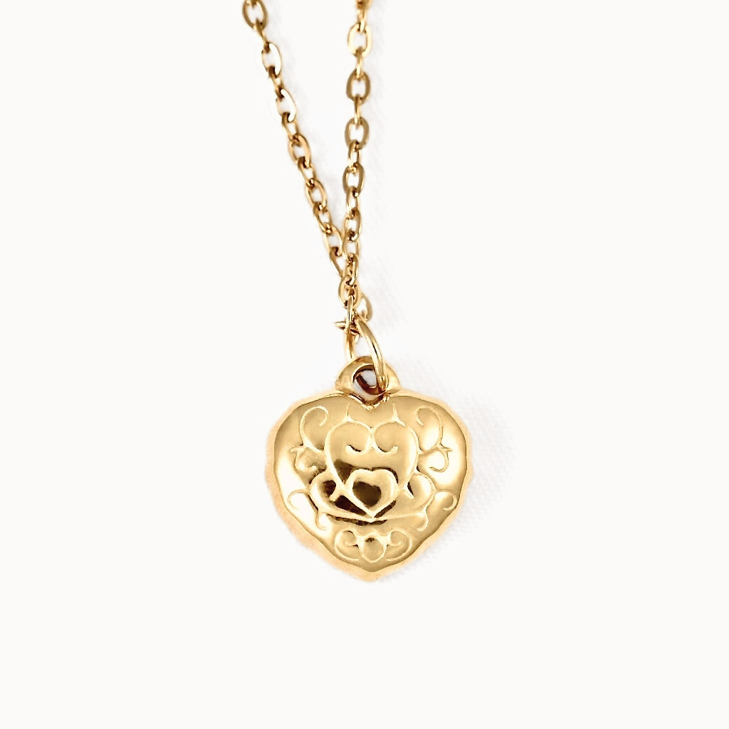 A gold heart-shaped pendant necklace with an intricate engraved design and a small heart in the center, hanging from a delicate gold chain against a plain white background.