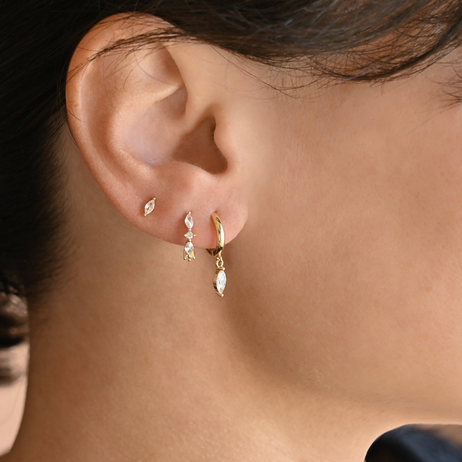 Close up of an ear wearing a gold stud earring with a single marquise stone, a huggie  hoop earring adorned with alternating marquise and round clear CZ stones and a another huggie hoop earring with a dangling marquise stone.