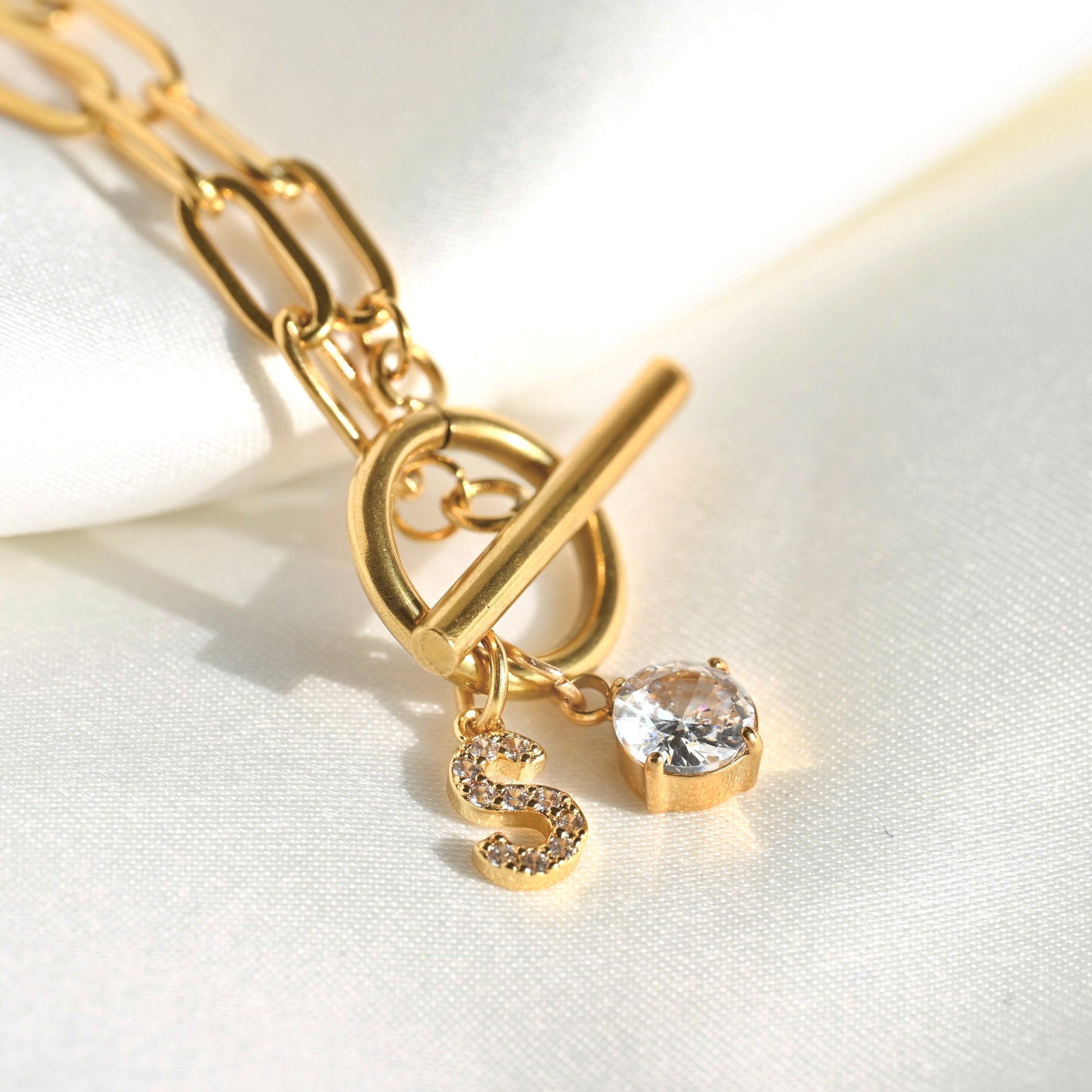 Gold toggle clasp chain bracelet with a dangling letter S charm and a clear heart-shaped crystal, shown on a white satin background.