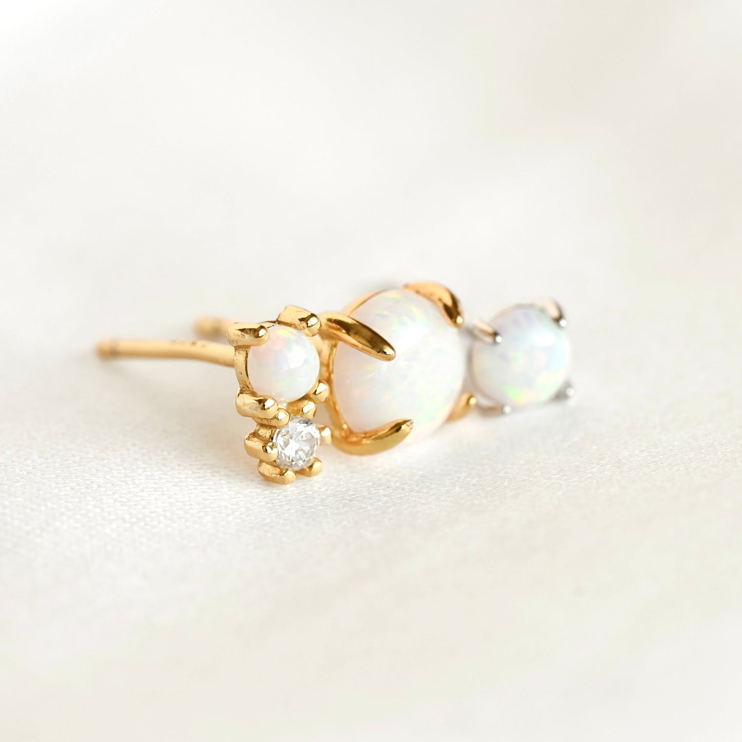 Two gold and one silver stud earring with opal stones of varying sizes and a small clear accent stone, photographed on a soft white background.
