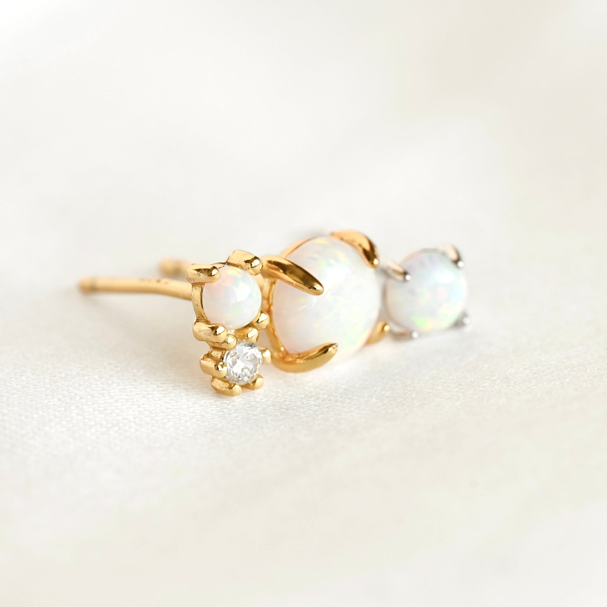 Two gold and one silver stud earring with opal stones of varying sizes and a small clear accent stone, photographed on a soft white background.